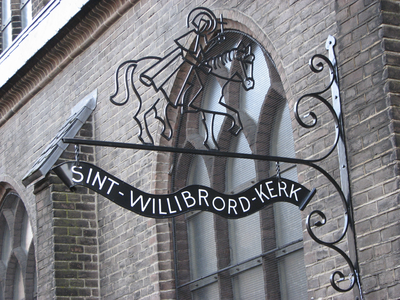 902872 Afbeelding van het smeedijzeren uithangteken 'SINT-WILLIBRORD-KERK' naast de ingang van de kerk ...